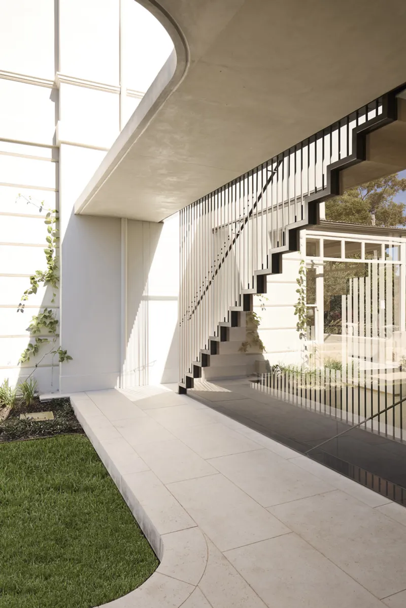 Concrete staircase with black metal railing beneath a curved soffit in a sunlit courtyard
