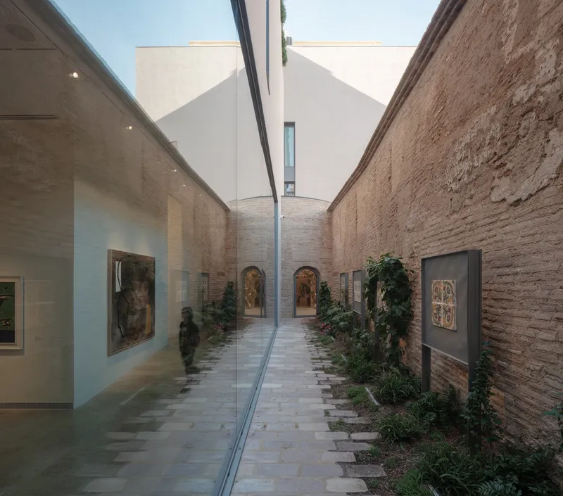 Narrow courtyard gallery with glass partition separating exhibited paintings from planted beds along exposed brick wall