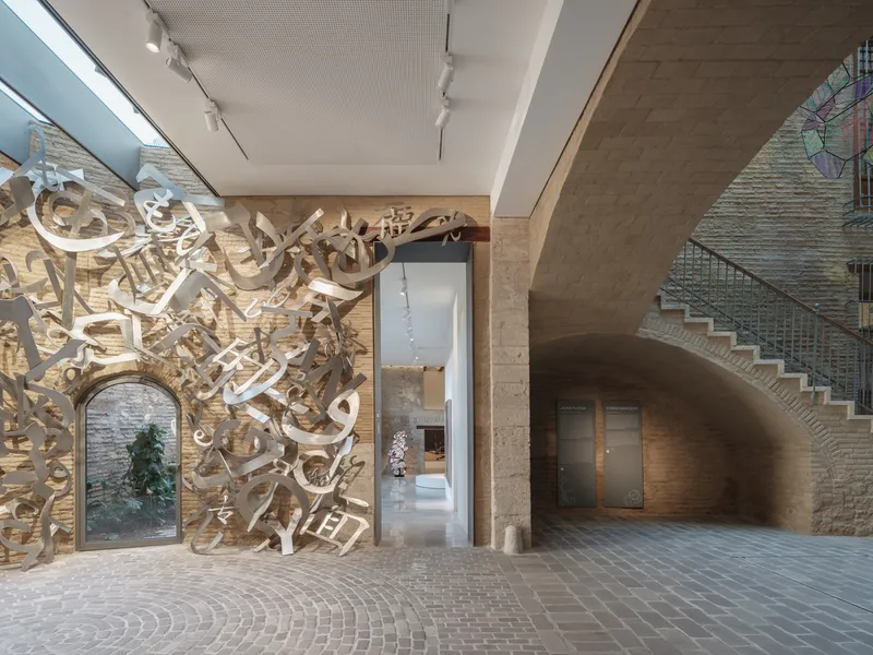 Brick-paved courtyard with sculptural installation against an arched stone wall and metal staircase