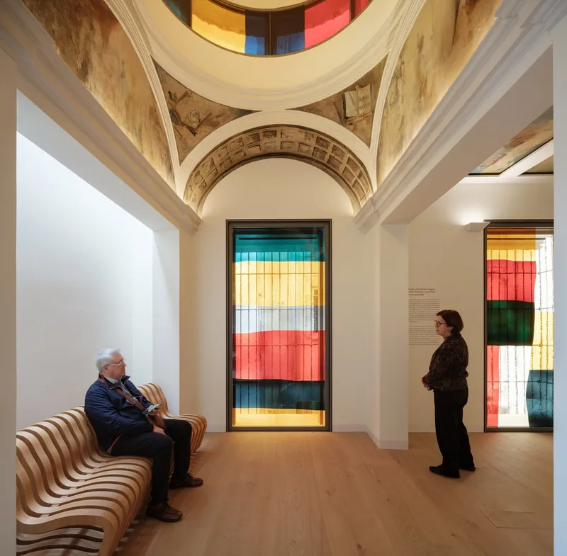 Gallery room with vaulted ceiling, ribbed wooden bench and two visitors viewing striped translucent panels
