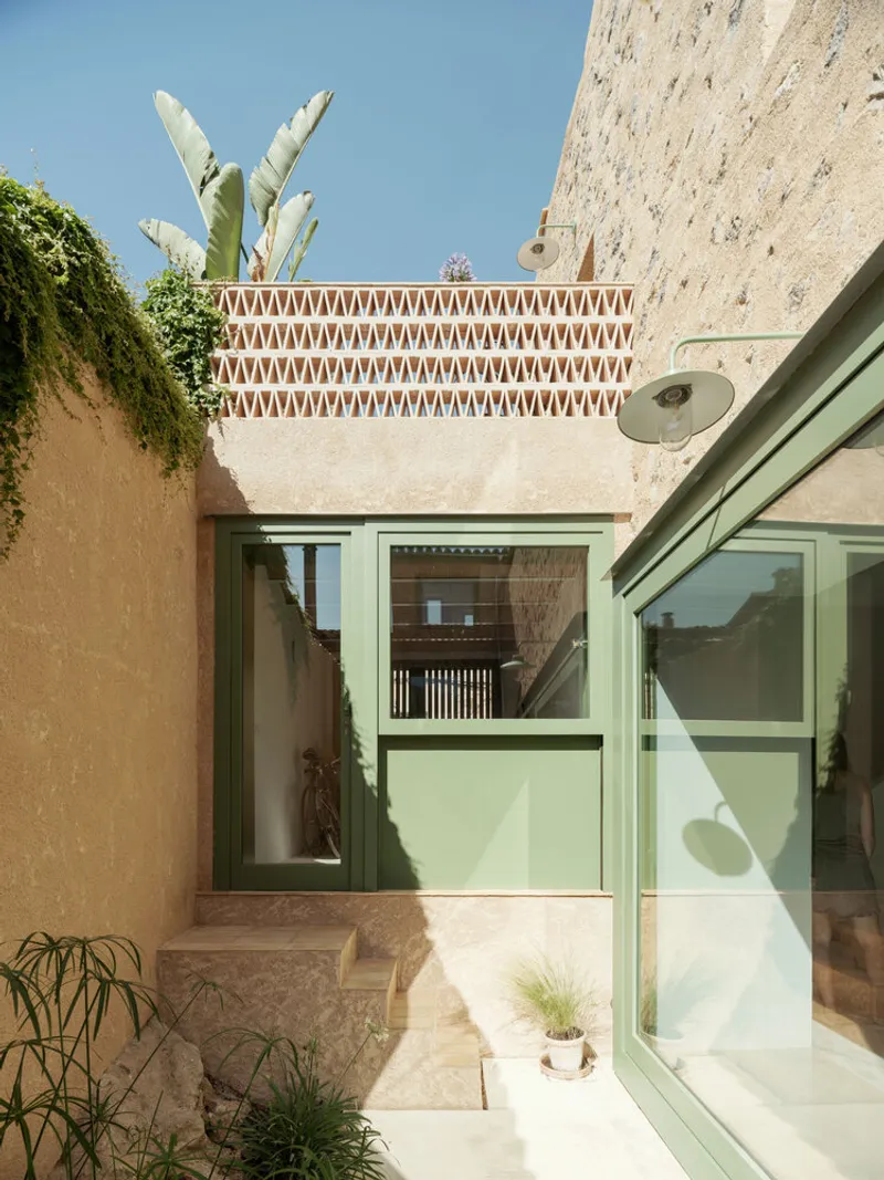 Exterior courtyard with green framed glazing beneath a terracotta patterned parapet and banana plant above stone walls