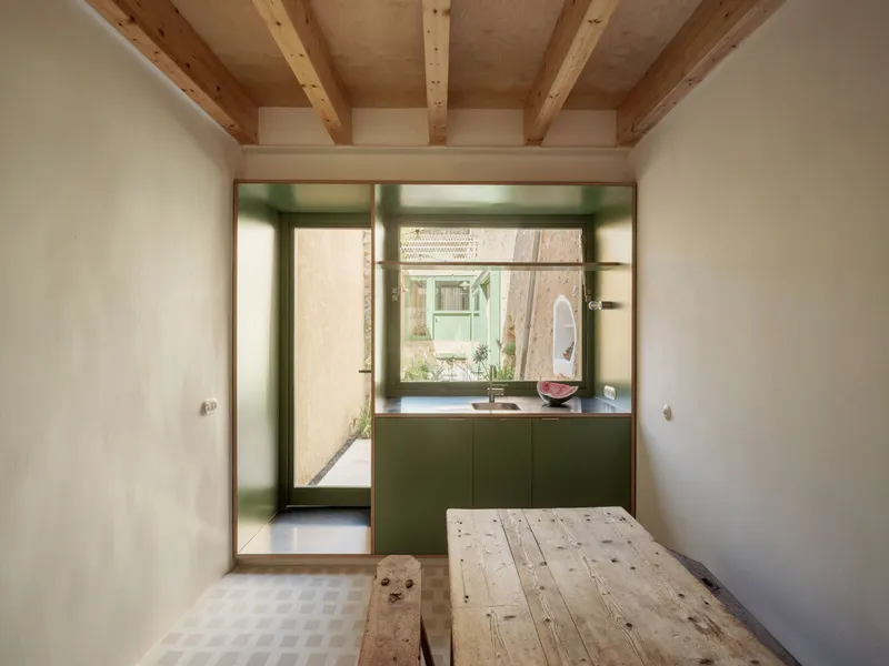 Green kitchen unit with integrated sink framed by timber opening beneath exposed ceiling joists and checkered floor