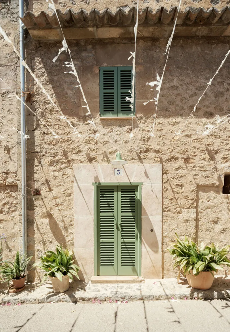Street facade with pale green louvered shutters and terracotta cornice detail under dappled tree shadows