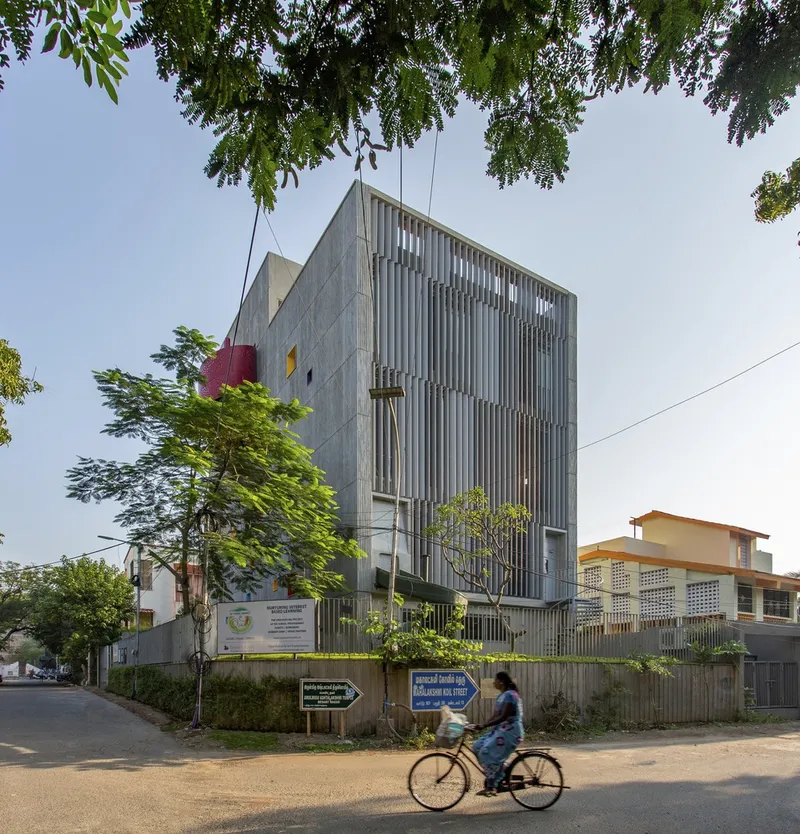 Street view of the concrete facade with vertical metal louvers framed by overhanging tree branches and a passing cyclist