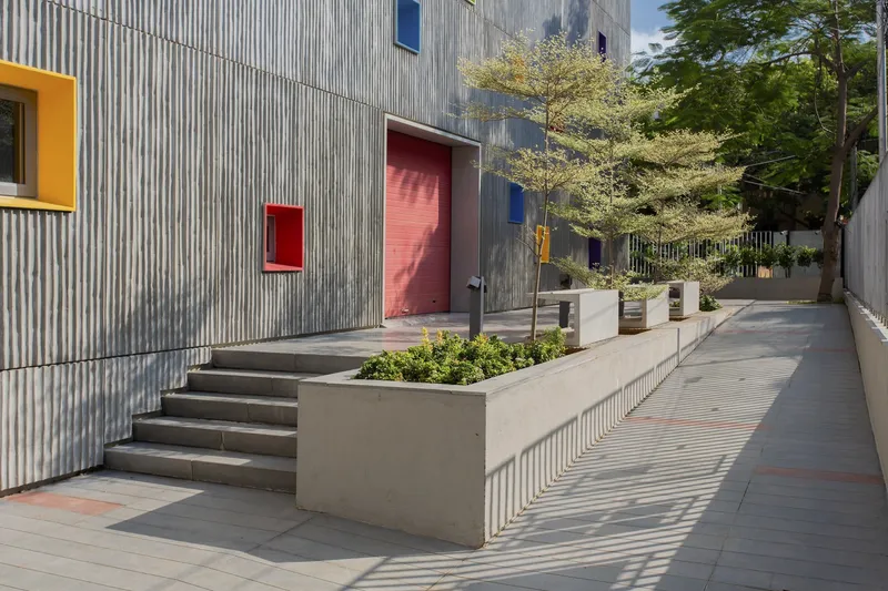 Exterior courtyard with concrete planters, metal steps, corrugated facade with colored door and window openings, and dappled sunlight
