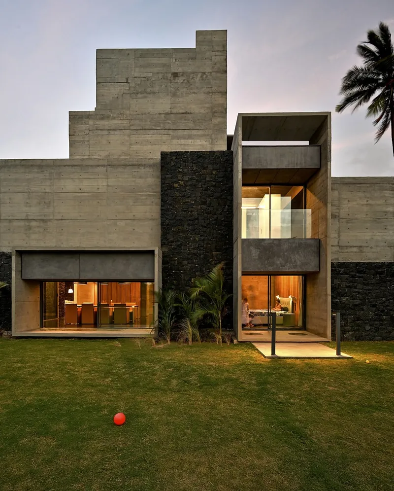 Board-formed concrete facade with stacked volumes and illuminated interiors at dusk beside a palm tree