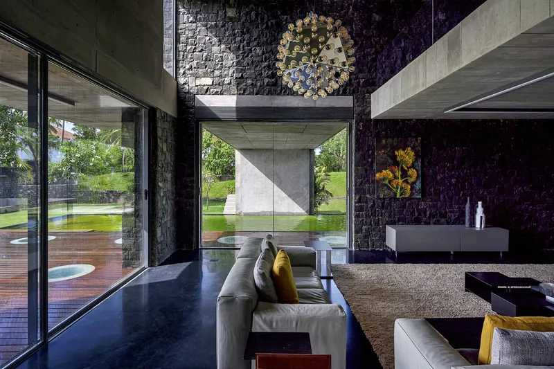 Double-height living space with textured dark stone walls and floor-to-ceiling glazing overlooking a reflecting pool