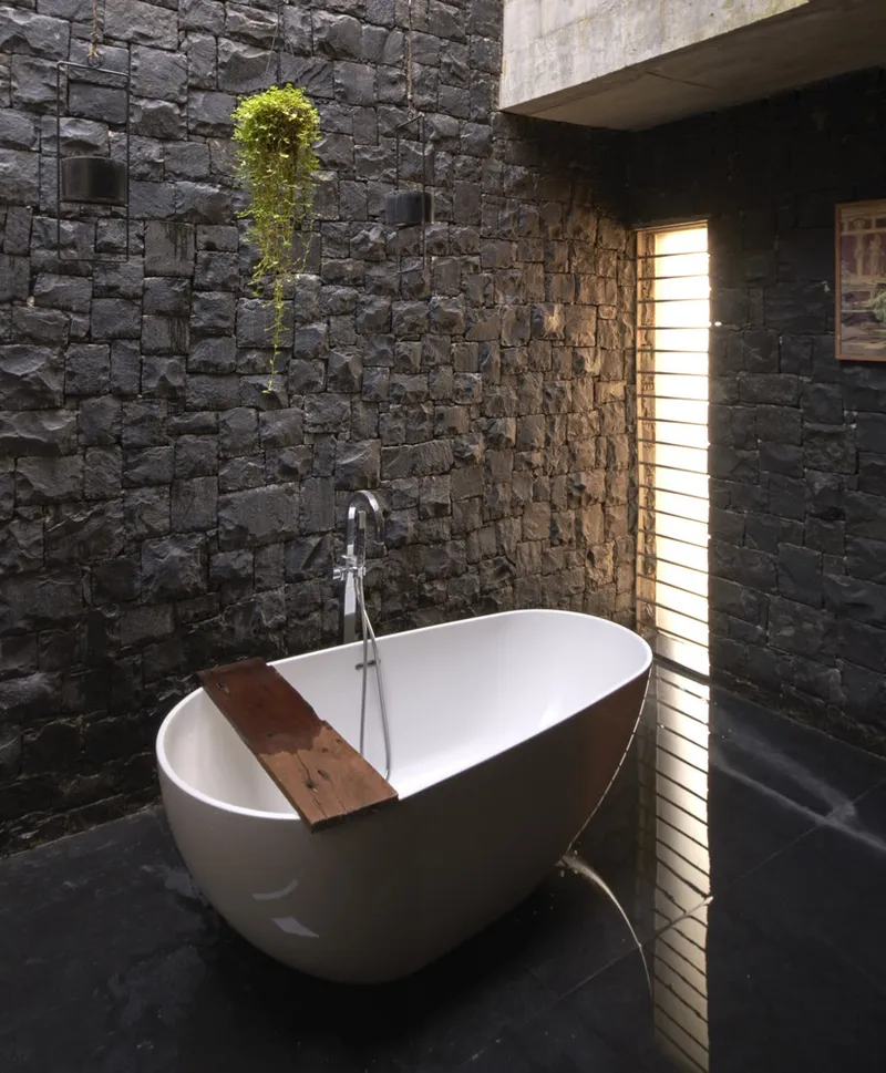 Freestanding white bathtub with timber tray beside dark stone wall with vertical slot window and plant