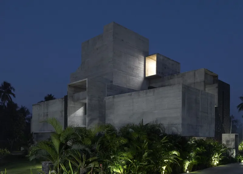 Stacked concrete volumes illuminated at dusk with palms and landscaped beds in foreground