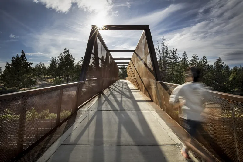 Steel walkway with perforated side panels casting geometric shadows as a cyclist passes through afternoon sun