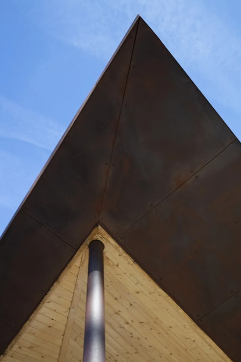 Upward view of the gabled roof corner with weathered steel cladding meeting a timber soffit and metal downspout