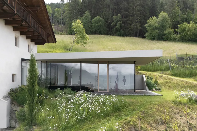 Glass-walled pavilion with flat concrete roof set into a sloping meadow with wildflowers