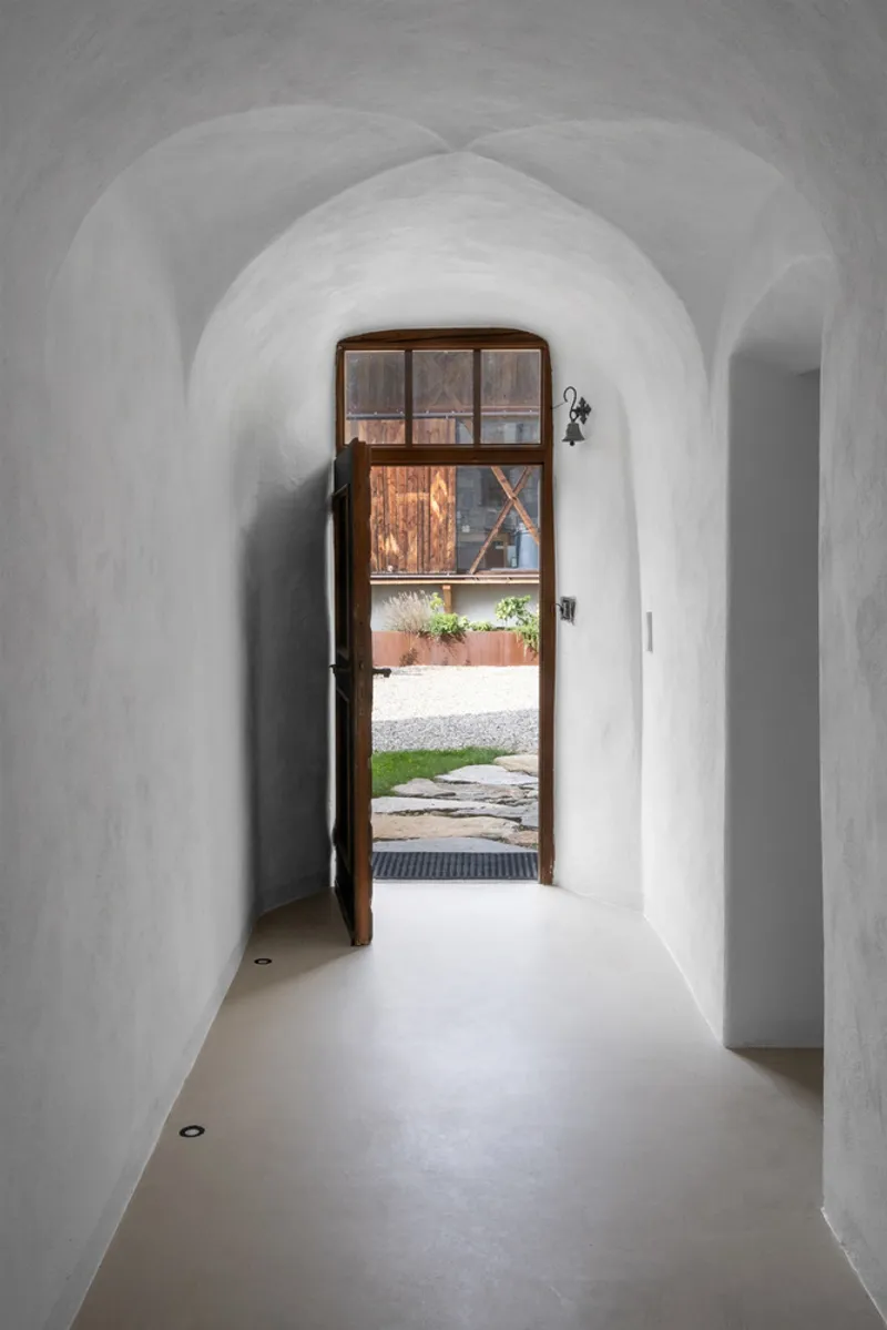 Vaulted white plaster corridor with timber and glass door open to a gravel courtyard