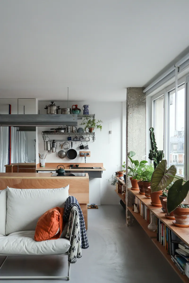 Low bookshelf with potted plants below exposed concrete column and aluminum window frames overlooking neighboring rooftops