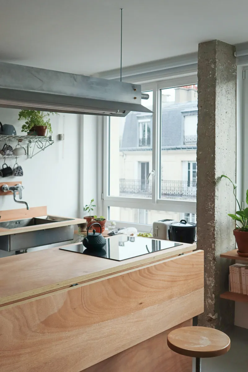 Plywood kitchen island with inset cooktop framed by exposed concrete column and galvanized steel ventilation hood