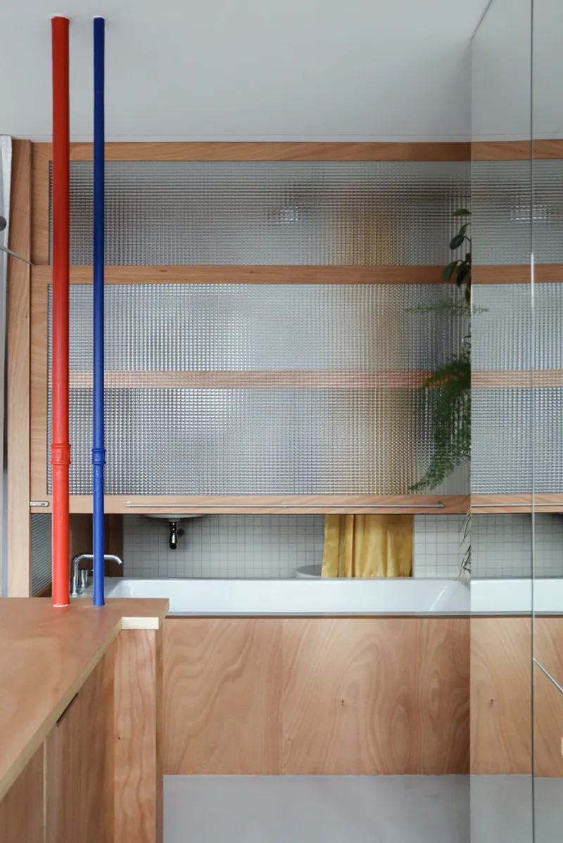 Plywood cabinetry with translucent reeded glass panels framed by horizontal timber strips and red and blue painted pipes