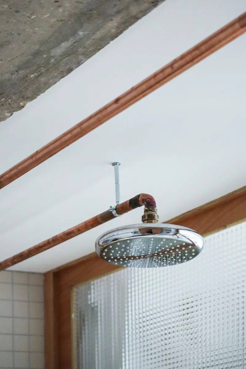 Chrome showerhead suspended from exposed copper plumbing against a white ceiling with timber beams