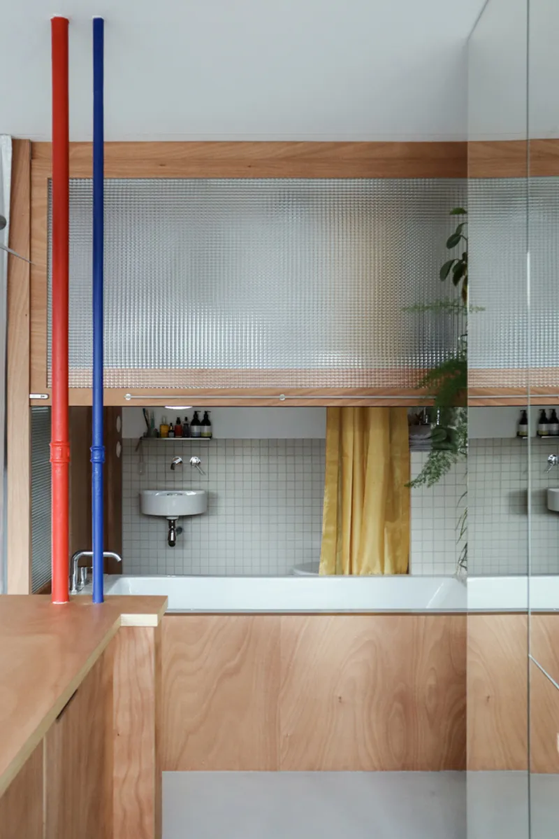 Split-level bathroom with translucent glass partitions, exposed red and blue pipes, and timber cabinetry