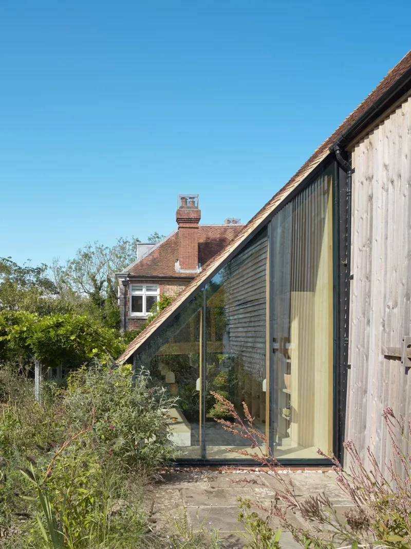 Glass extension set into the sloped roof of a weathered timber barn with planted edges