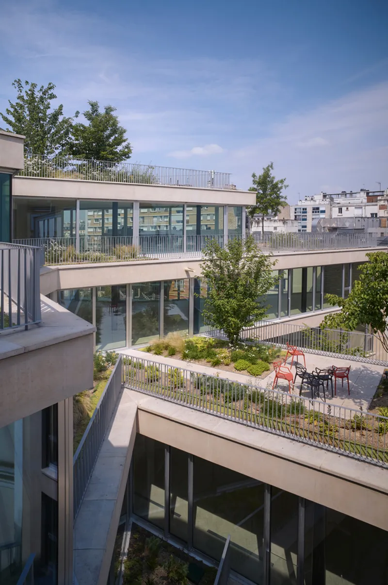 Terraced courtyard with planted green roofs, metal railings and red cafe chairs under a clear sky