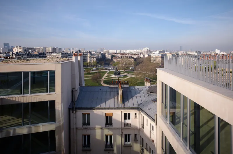 Rooftop view across zinc-clad volumes toward a public park under clear afternoon sky