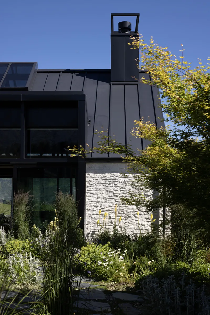 Zinc-clad chimney rising above a black metal roof with white brick base and garden planting below