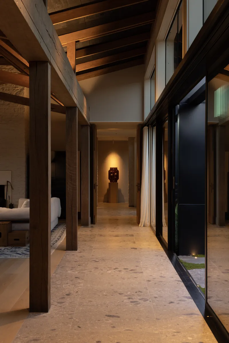 Corridor with exposed timber columns and ceiling beams leading toward a backlit sculpture niche