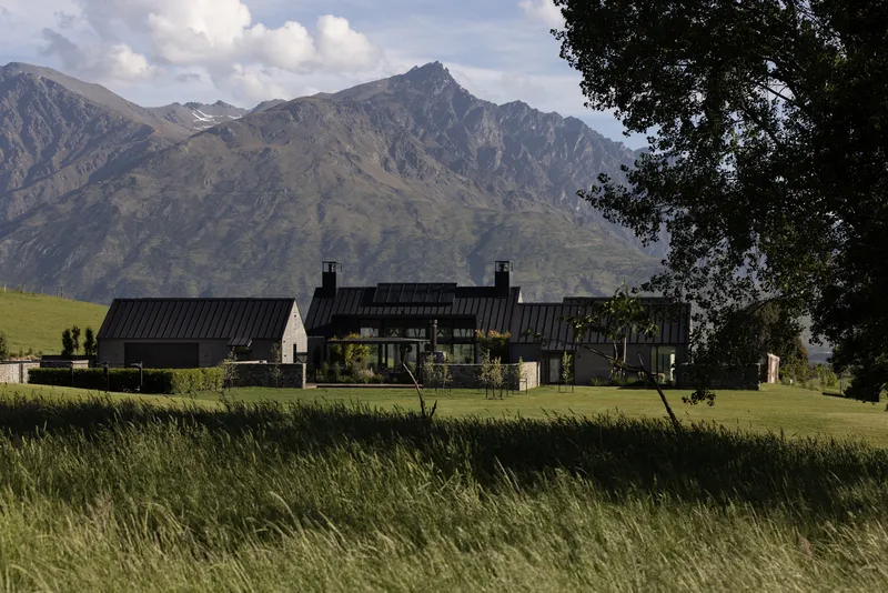 Group of black-roofed volumes set in a meadow with mountain peaks in the distance