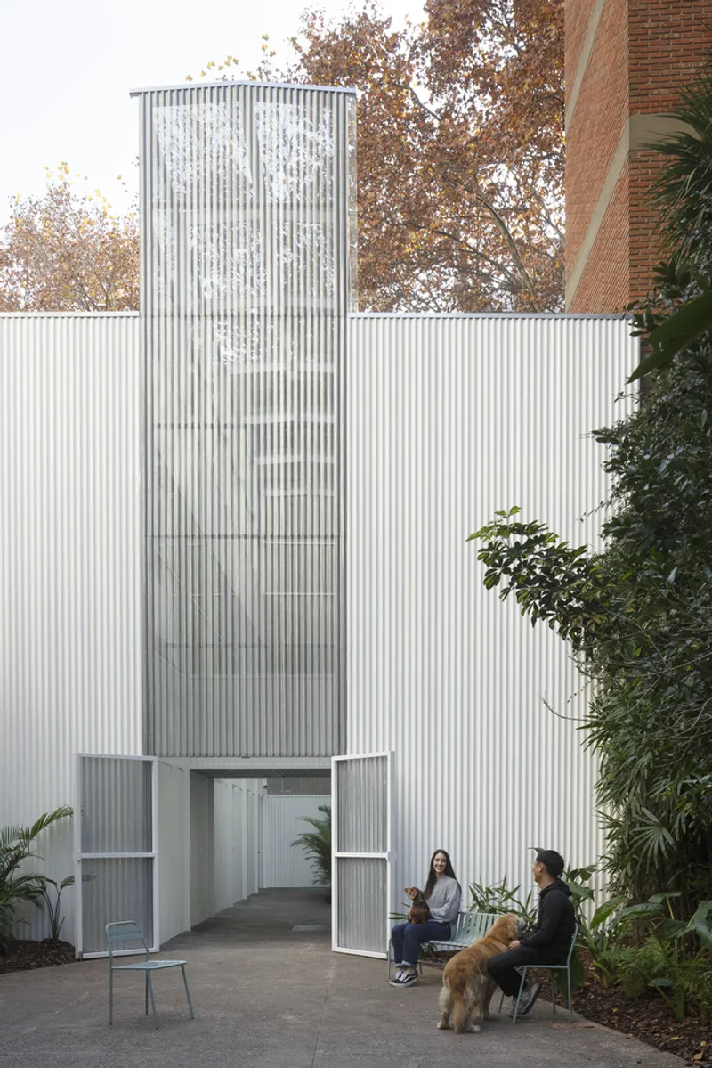 Entrance courtyard with corrugated metal walls and cylindrical volume where two people sit with a dog
