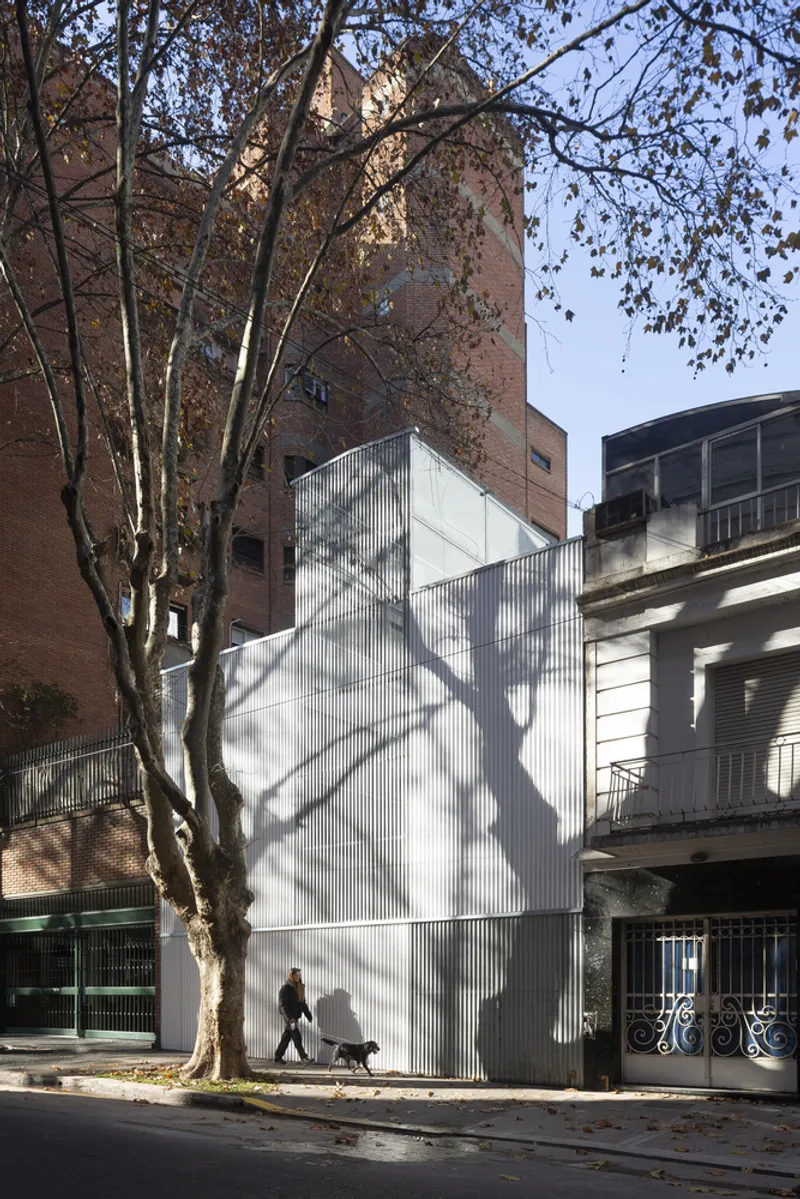 Narrow facade with vertical corrugated cladding casting tree branch shadows as a pedestrian passes by