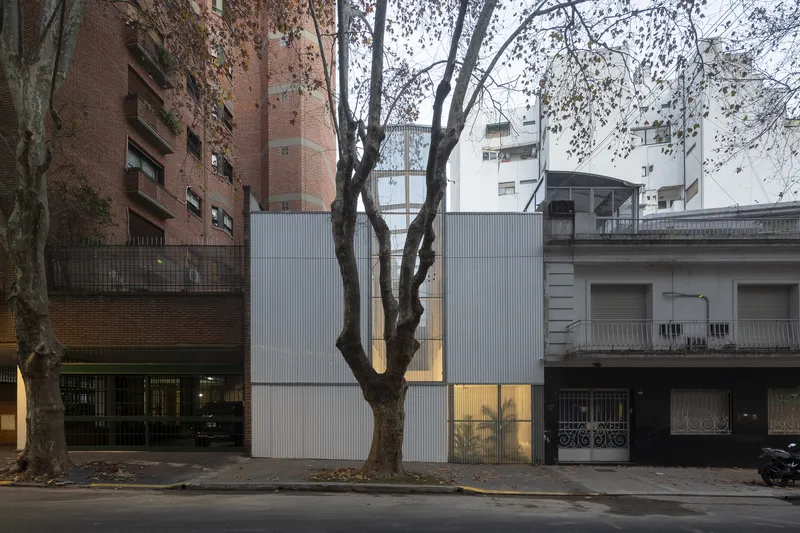 Street view of the corrugated metal facade wedged between apartment buildings with a mature tree in front