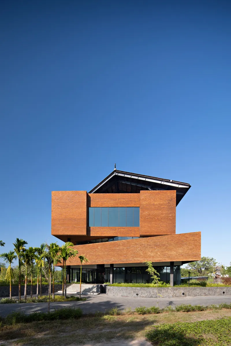 Stacked orange brick facade with projecting volumes above a stone base wall and flanking palm trees