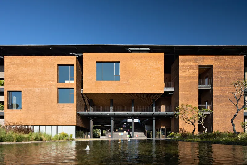 Rammed earth facade volumes raised on pilotis above a reflecting pond under a clear blue sky