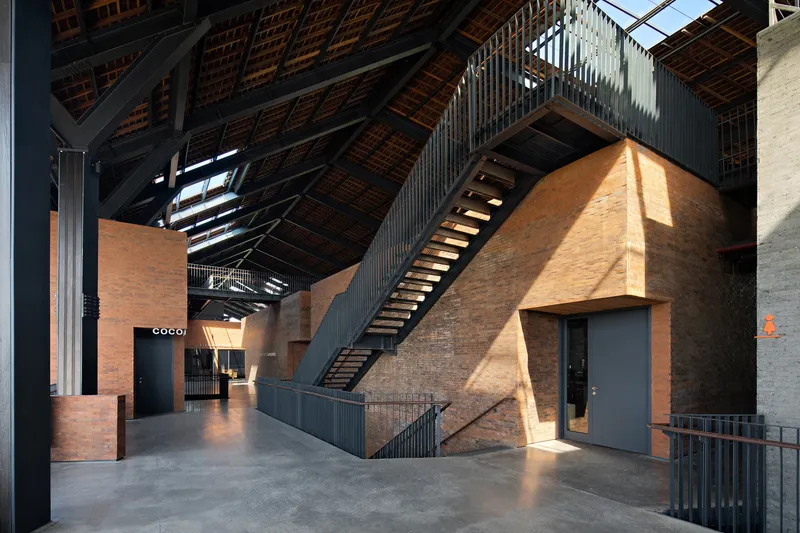 Interior atrium with steel staircase ascending along a rammed earth wall beneath an exposed timber roof structure