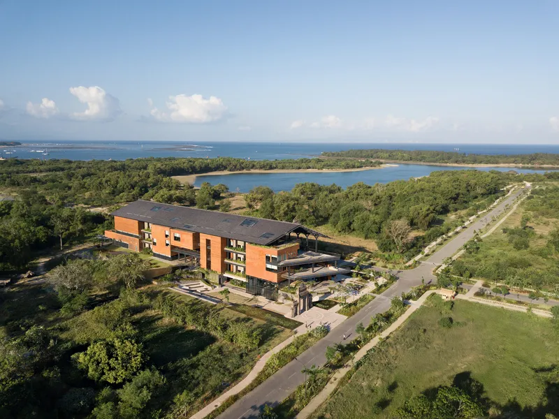 Aerial view of the multi-story brick and terracotta volume set among trees with coastal lagoon beyond