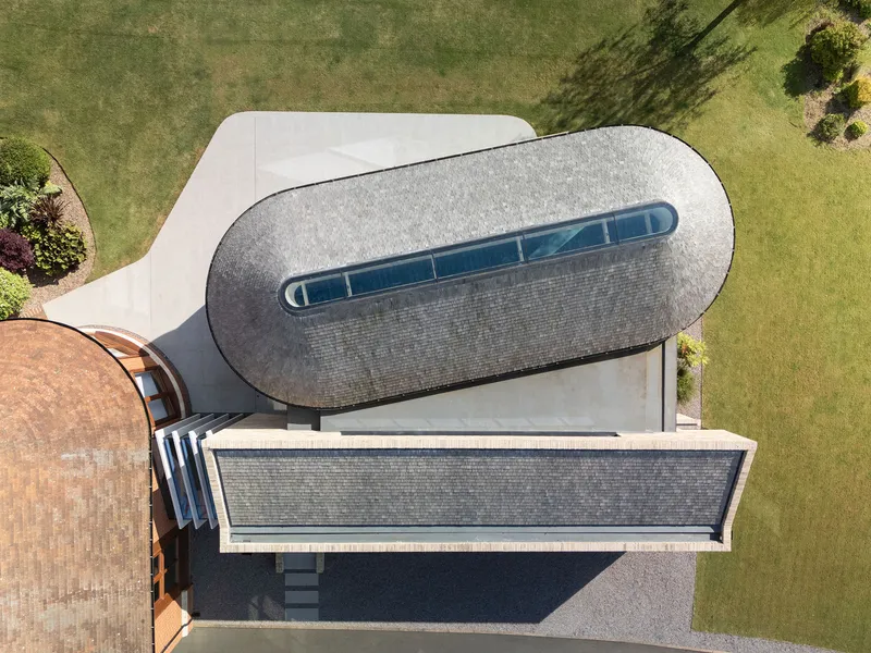 Aerial view of the oval-shaped shingled roof with a linear skylight surrounded by green lawns