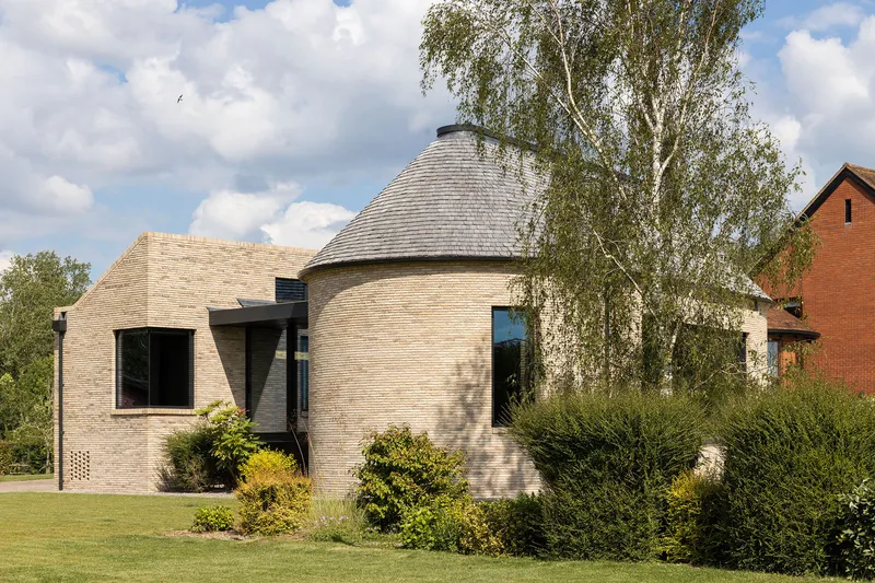 Buff brick facade with cylindrical shingled roof volume framed by birch trees and planted garden beds