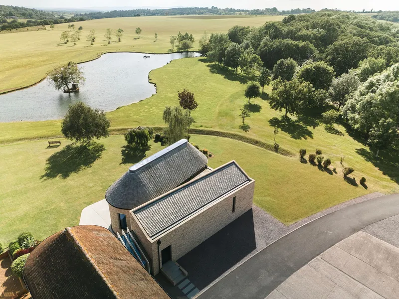 Drone view showing the brick volumes beside a curving pond and mature tree groupings