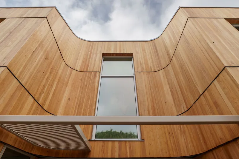 Upward view of the undulating timber facade with a vertical window slot beneath cloudy sky