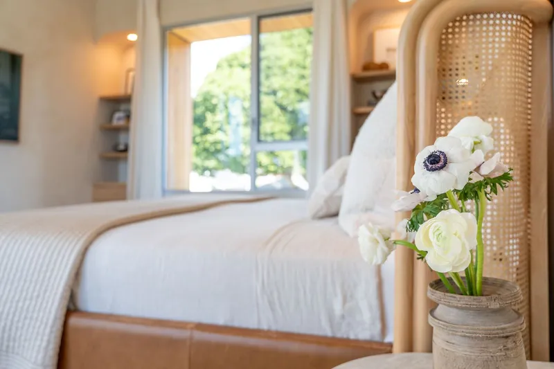 Bedroom corner with cane headboard and vase of white flowers beside a window overlooking green trees
