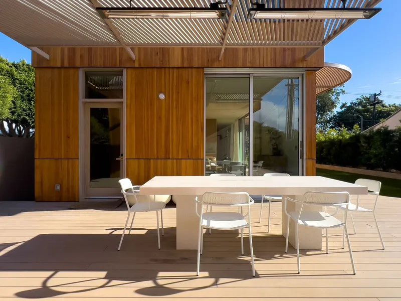 Timber-clad rear facade with slatted pergola overhang and deck dining area under blue sky