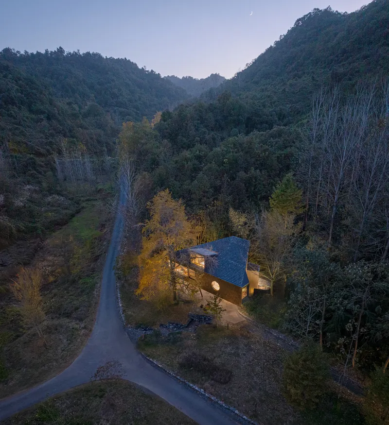 Aerial view of the house nestled among forested hills at dusk with warm interior lighting