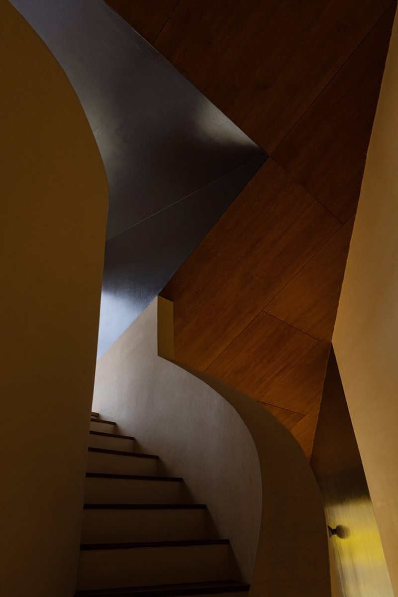 Curved stairwell with intersecting plaster and timber ceiling planes under directional natural light