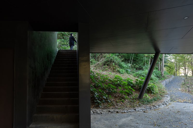 Interior staircase ascending to a silhouetted figure with an opening framing the wooded hillside