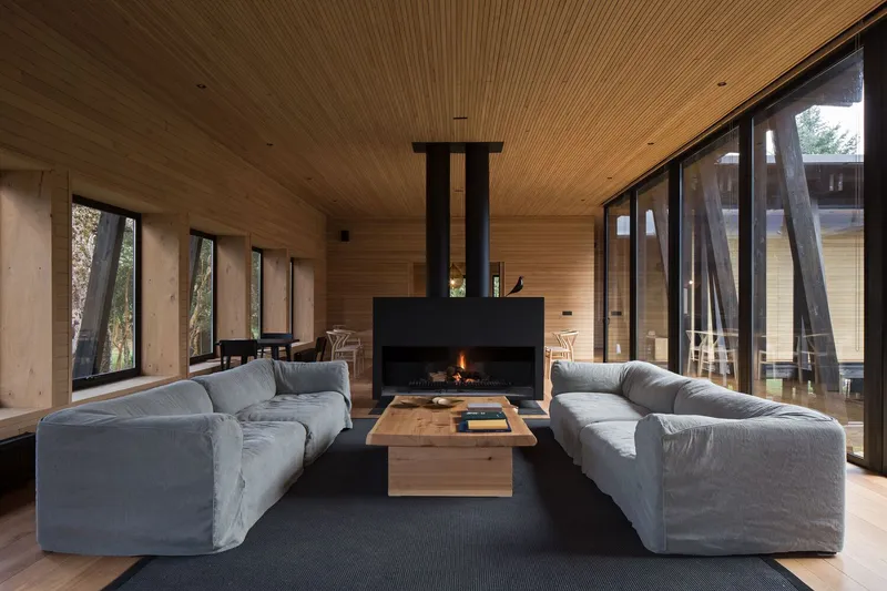 Living room with wood-lined ceiling and walls, a central fireplace, and glazed doors opening to the forest