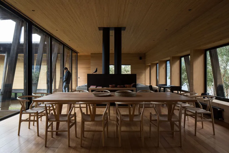 Open living and dining space with long timber table, angled glazing and central fireplace with figure passing by