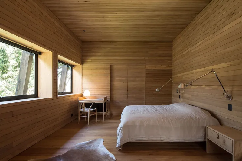 Timber-lined bedroom with horizontal plank walls and ceiling, framed windows looking onto green foliage