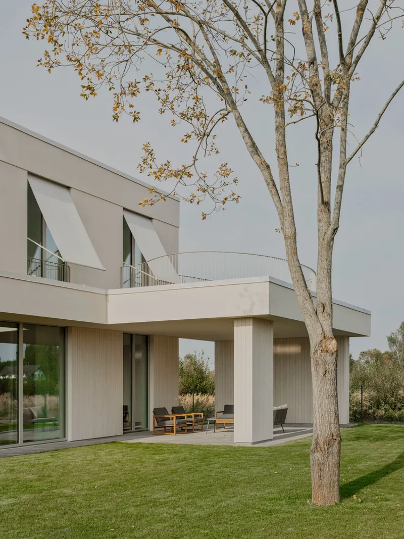 Garden view showing covered terrace with white columns under upper-level balcony beside deciduous tree