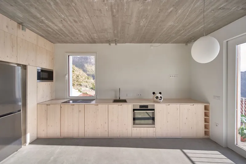 Light wood kitchen cabinetry against white walls beneath a board-formed concrete ceiling in afternoon shadow