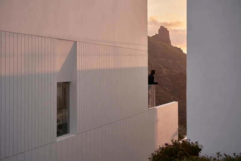 Corrugated white volume with balcony and silhouetted figure against a rocky peak at sunset